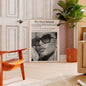 Living room with a framed newspaper featuring a woman's face, orange chair, and plant.